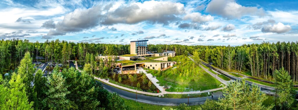 The Kurro Spa complex in the middle of a pine forest: green roofs, timber facades and a glass tower under cumulus clouds, drone panorama
