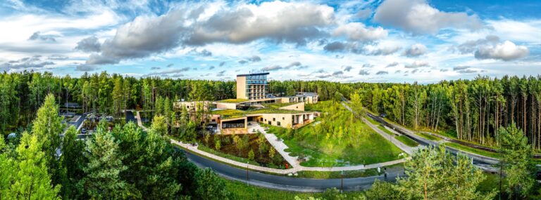 The Kurro Spa complex in the middle of a pine forest: green roofs, timber facades and a glass tower under cumulus clouds, drone panorama