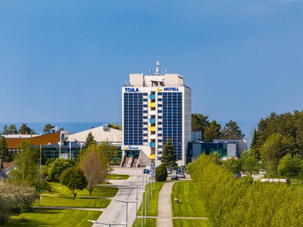 The tall main building of Toila Spa Hotel from the front with the approach road and spring greenery, the Gulf of Finland behind, drone view