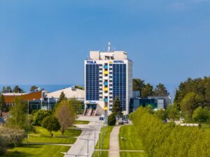 Высокое главное здание отеля Toila Spa спереди, подъездная дорога и весенняя зелень, позади Финский залив, вид с дрона