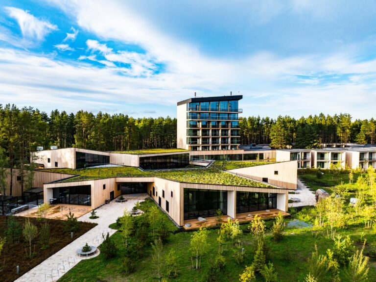 The low timber-clad wings of Kurro Spa with green roofs and the glass tower behind them, a paved footpath in front, drone view