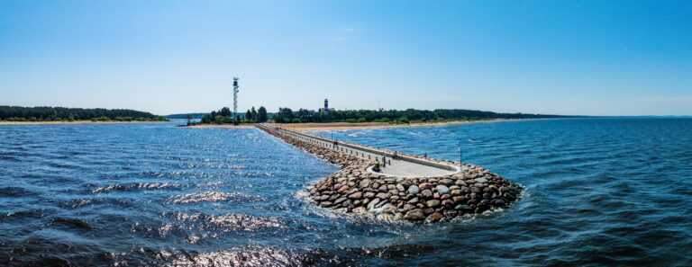 Мол Нарва-Йыэсуу сбоку с малой высоты над водой, синее море и небо, позади берег, панорама с дрона