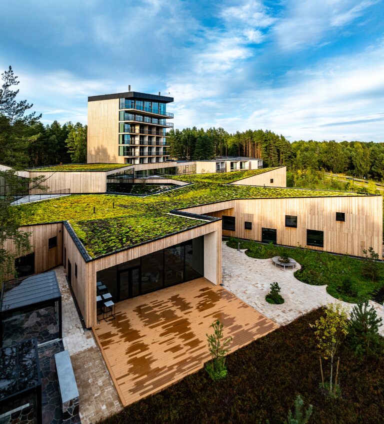 A terrace and green roof of Kurro Spa in the foreground, the glass tower and pine forest behind, vertical drone panorama