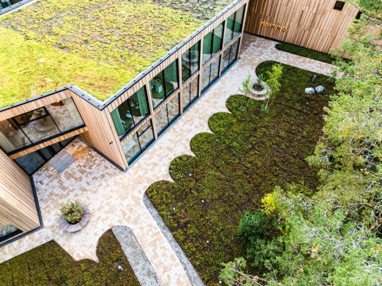 The timber facade and glass walls of Kurro Spa from above, beside a paved courtyard with a scalloped planting bed and a green roof