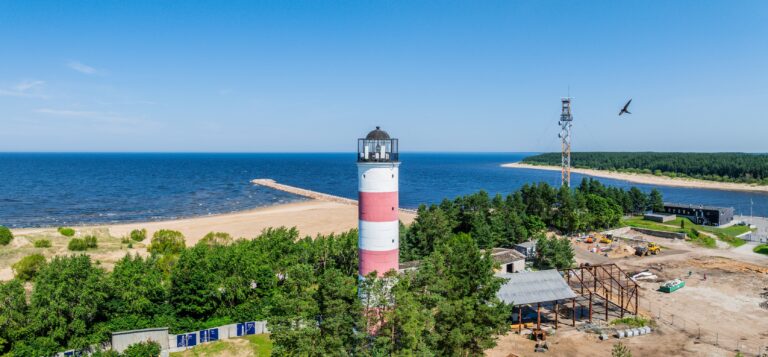The red-and-white Narva-Jõesuu lighthouse among the trees with a construction site beside it, the beach, breakwater and sea behind, drone panorama
