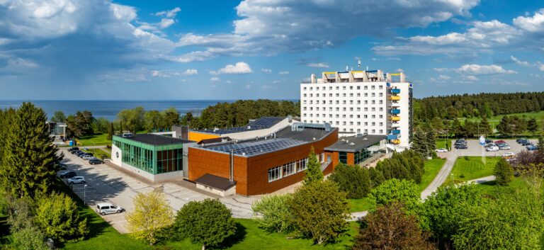 Крыло спа Toila Spa с коричневой обшивкой и белое здание отеля, позади лес и море, панорама с дрона
