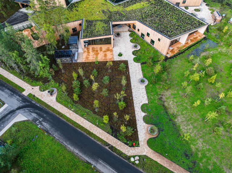 A green-roofed wing of Kurro Spa with timber terraces from above, a bed of young trees and a paved path beside it