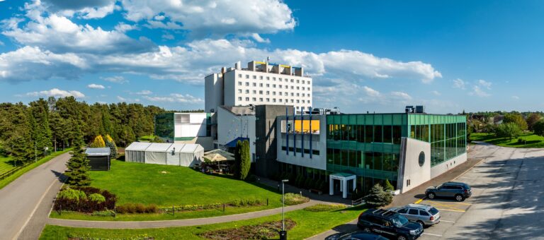 Здание Toila Spa с зелёным стеклянным фасадом, за ним главное здание отеля, впереди дорога и парковка, панорама с дрона
