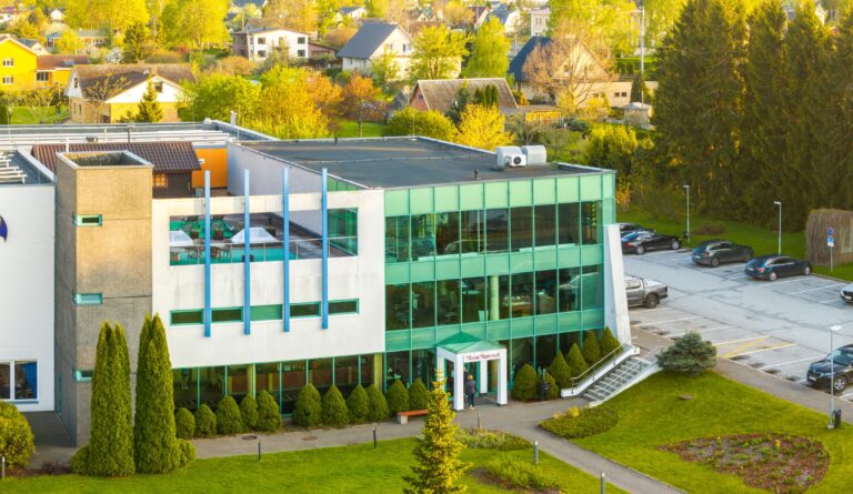 Здание Toila Spa с зелёным стеклянным фасадом и терраса на крыше в вечернем свете, позади дома посёлка, вид с дрона