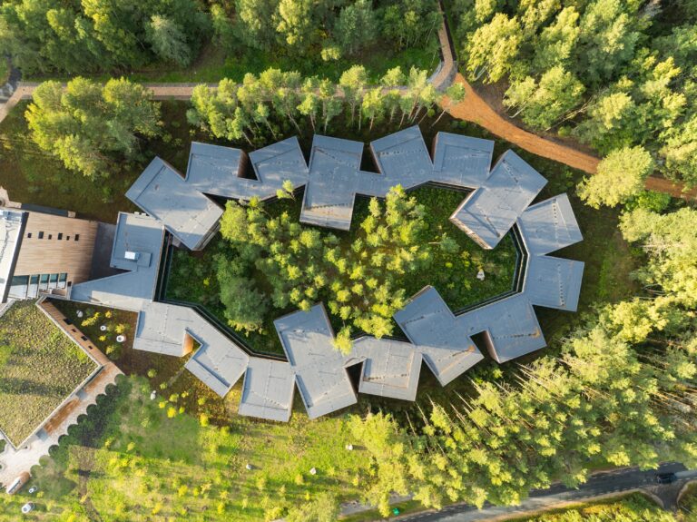 The ring-shaped wing of Kurro Spa around a pine grove from directly above, grey roofs set at angles, drone view