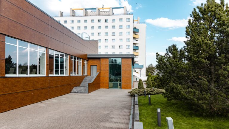 Крыло Toila Spa с коричневой обшивкой и большими окнами, позади главное здание отеля, рядом сосна